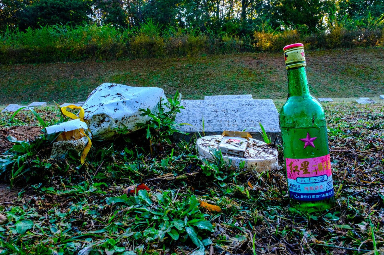 Offered by an unknown admirer, a bottle of Chinese liquor, a pack of cigarettes (also Chinese) and a bouquet of faded flowers honour the grave of an anonymous North Korean soldier (October 2024).