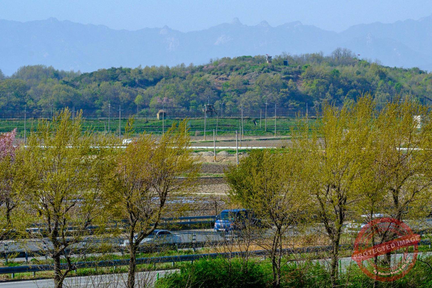 jayu-ro-north-korea-border Cars drive along Jayu-ro with a South Korean surveillance tower in the background and North Korean guard posts with their flags at the top of the hill (April 2025).